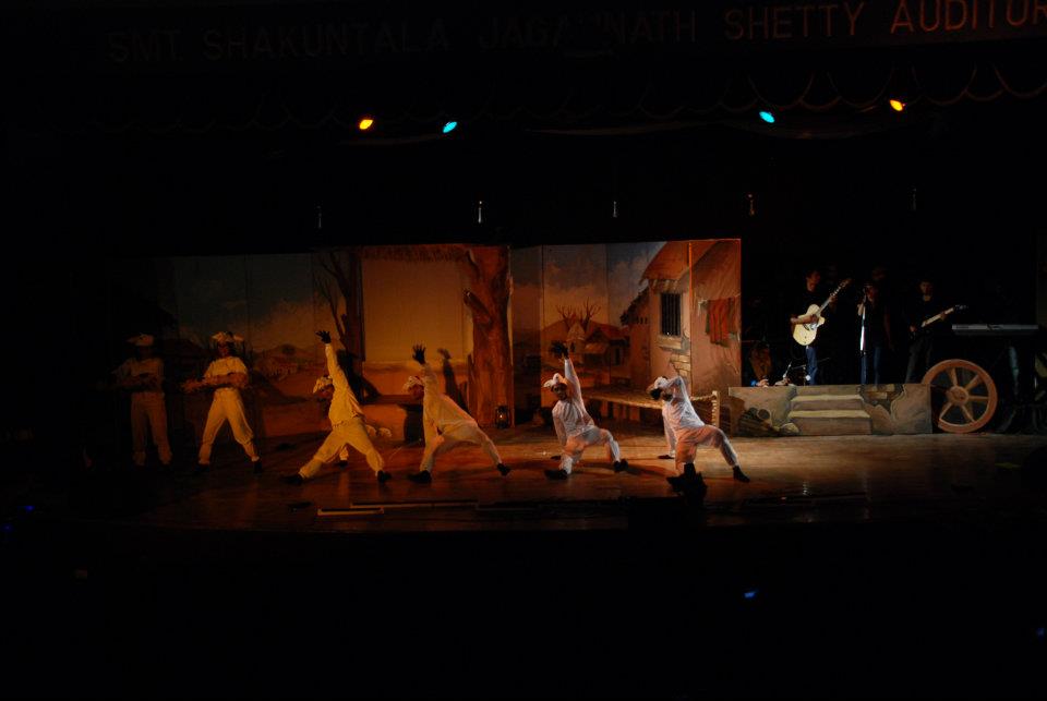 Two masked sheep characters facing the seated farmer on stage